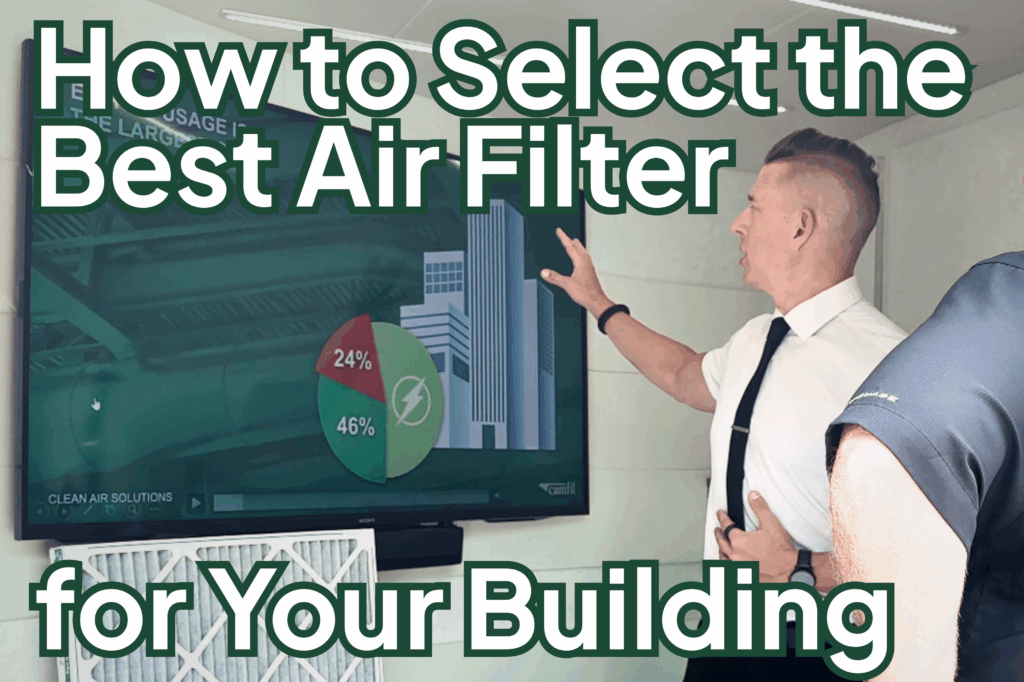 A man points at a large computer screen and explains energy cost savings in a building. He has a thin black tie and a short-sleeve white dress shirt and black pants. He has a fade with longer blonde hair on top. 
